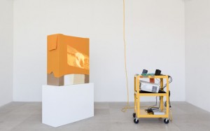 Rachel Harrison, "AA," 2010, wood, bubble wrap, cardboard, acrylic, tennis shirt, A/V cart, DVD player, speakers, projector, extension cord, five hair rollers, pack of gum, ear plugs, American Apparel video, color/sound (2009), 80 x 70 x 70 inches. Courtesy of Greene Naftali Gallery.